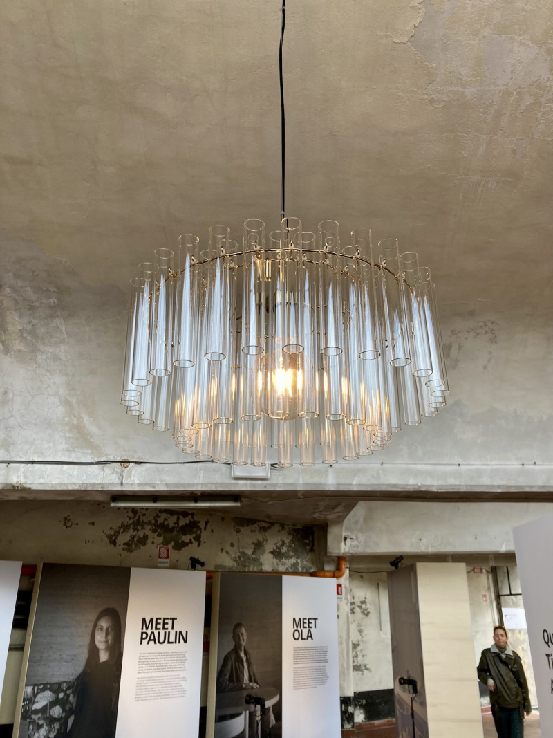 Fabrikhalle mit Betonwänden, von der Decke hängend eine Lampe, bestehend aus einem inneren und einem äußeren Ring, an dem viele kleine Glasröhrchen befestigt sind, im Raum, frontal, 2 Plakatwände und eine Person in einem Durchgang