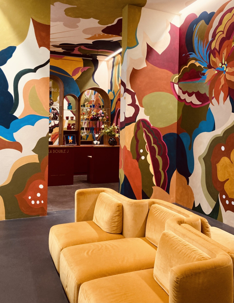 goldfarbenes Samtsofa mit Kissen und 6 Sitzmöglichkeiten, vor einer mit floralen Motiven bunt bemalten Wand und einer bordeaufarbenen Bar im Hintergrund