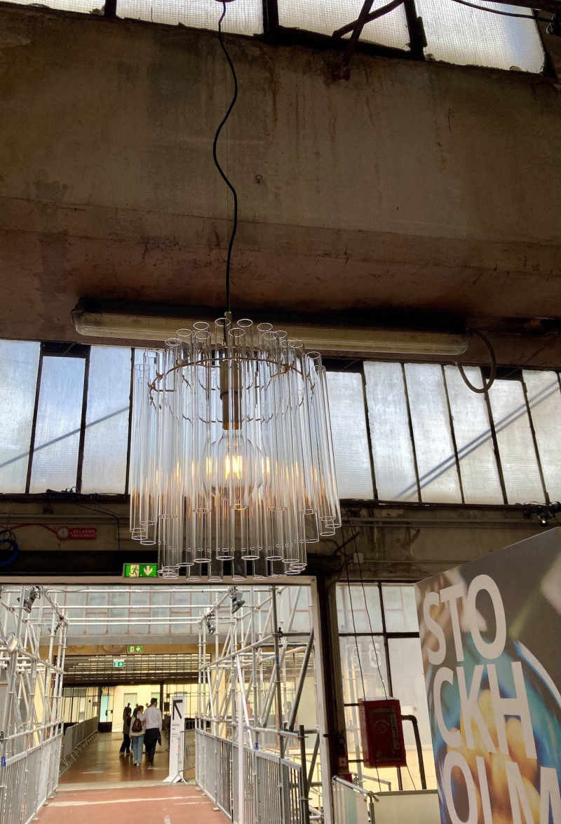 Fabrikhalle mit Betonwänden und vielen schmalen Glasfenstern, von der Decke hängend eine Lampe, bestehend aus einem inneren und einem äußeren Ring, an dem viele kleine Glasröhrchen befestigt sind, im Raum rechts eine Plakatwand, in der Mitte des Raums mehrere Personen in einem Durchgang