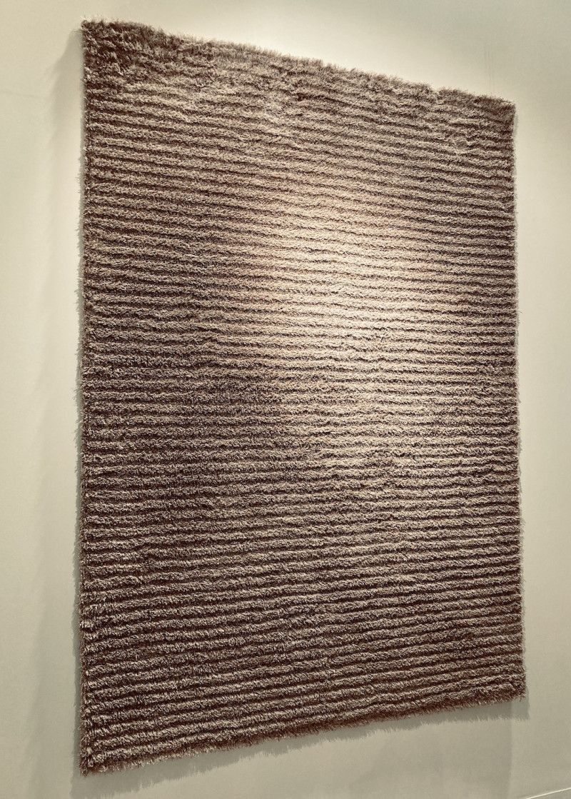 auf sandfarbener Wand hängt ein graubrauner, quadratischer Teppich, dessen Flor ein Streifenmuster erzeugt