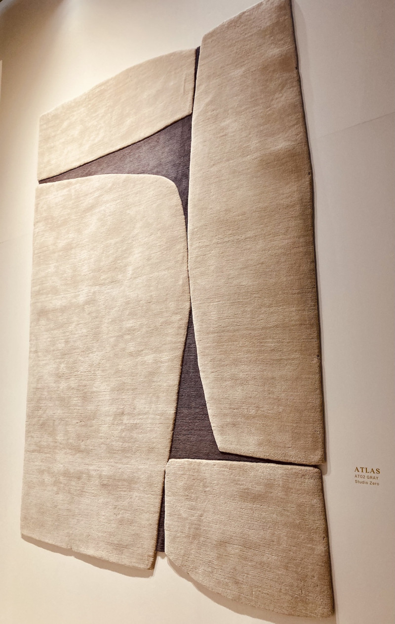 an einer sandfarbenen Wand hängt ein annähernd quadratischer, großer Teppich, bestehend aus 4 sandfarbenen Feldern, unterschiedlicher Größe, die Zwischenräume sind braun