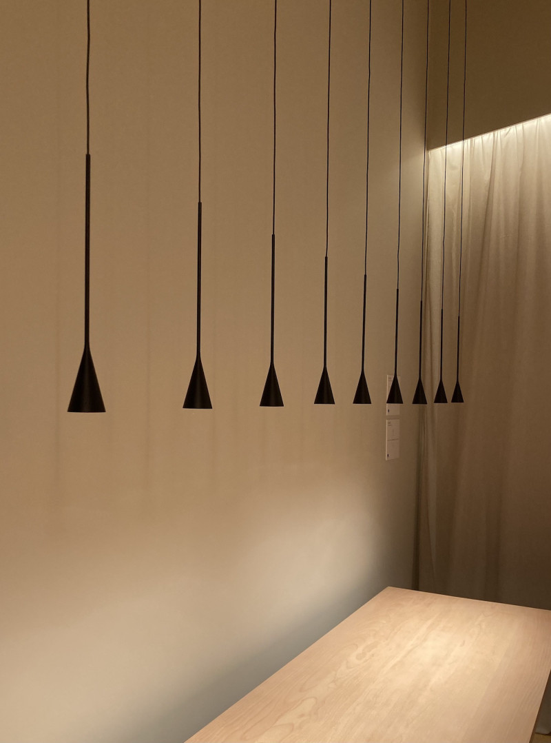 9 kleine, schwarze, konische Lampen, in Reihe gehängt über einem hellen Holztisch, an einer sandfarbenen Wand, am Ende des Tisches ein beleuchteter gleichfarbiger Vorhang