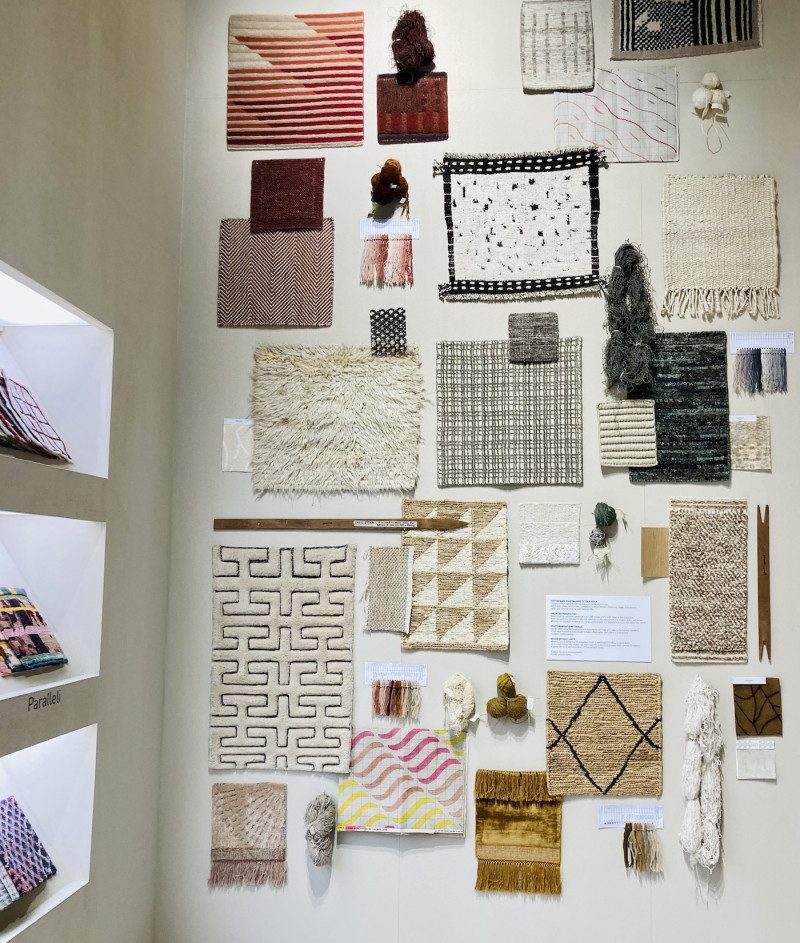 weiße Wand mit vielen verschiedenen Teppichmustern unterschiedlicher Farbe, auf einer großen Fläche quadratisch angeordnet, links im rechten Winkel, eine Wand mit beleuchteten Nischen, darin ebenfalls Teppichmuster