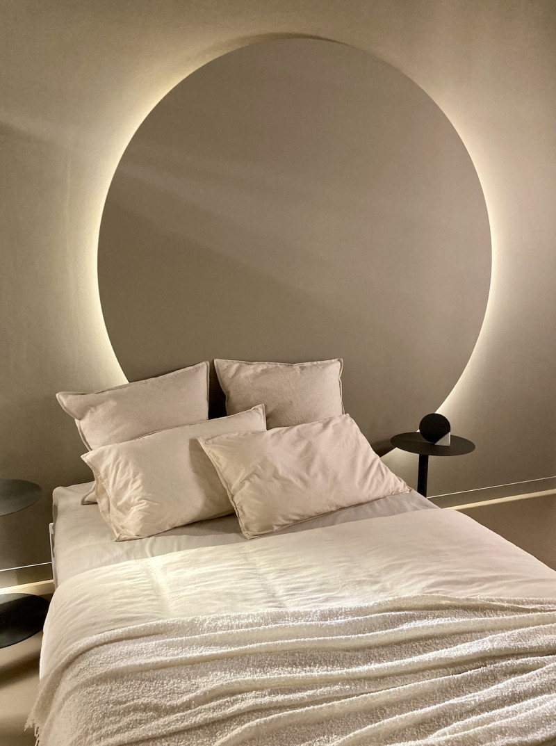 indirekte Beleuchtung hinter einer grauen Scheibe am Kopfteil eines Bettes, weiße Bettwäsche, 4 Kissen und 1 Decke, links und rechts vom Bett jeweils ein kleiner runder Nachttisch