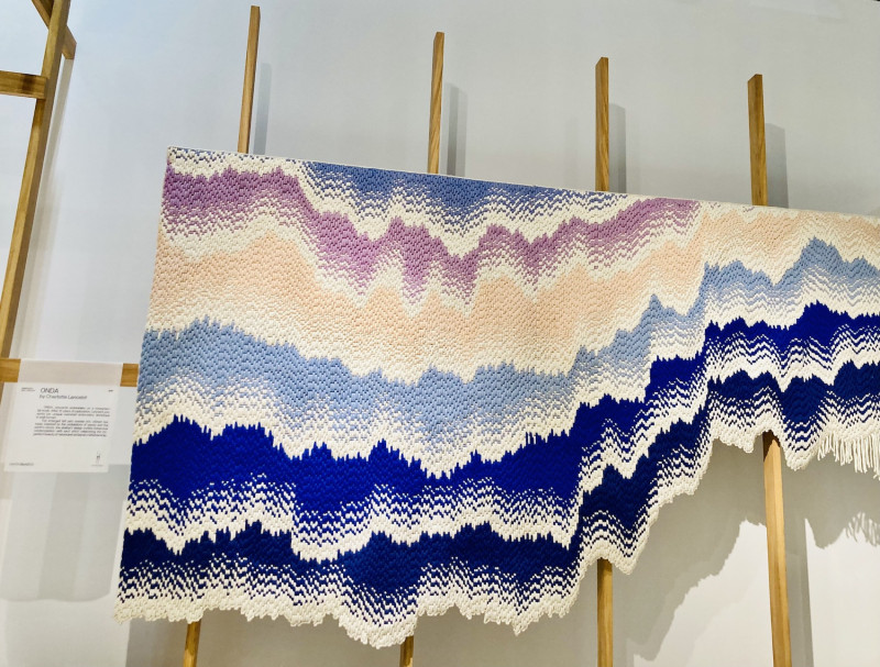 Holzgestänge, an dem ein Teppich befestigt ist, der ein Wellenmuster hat, Farben: Dunkelblau, Mittelblau, Weiß, Creme, Apricot und Lila, am unteren Rand verläuft die Webkante in kleinen und großen Wellen