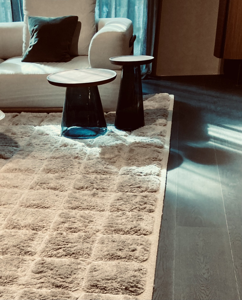 wollweißer rechteckiger Teppich mit hochflorigen Quadraten als Muster, liegt auf grauem Holzboden, auf dem Teppich 2 Beistelltische mit blauem Glasfuß und Holzplatte in unterschiedlicher Größe, dahinter weiße Couch mit weißem und grünen Kissen