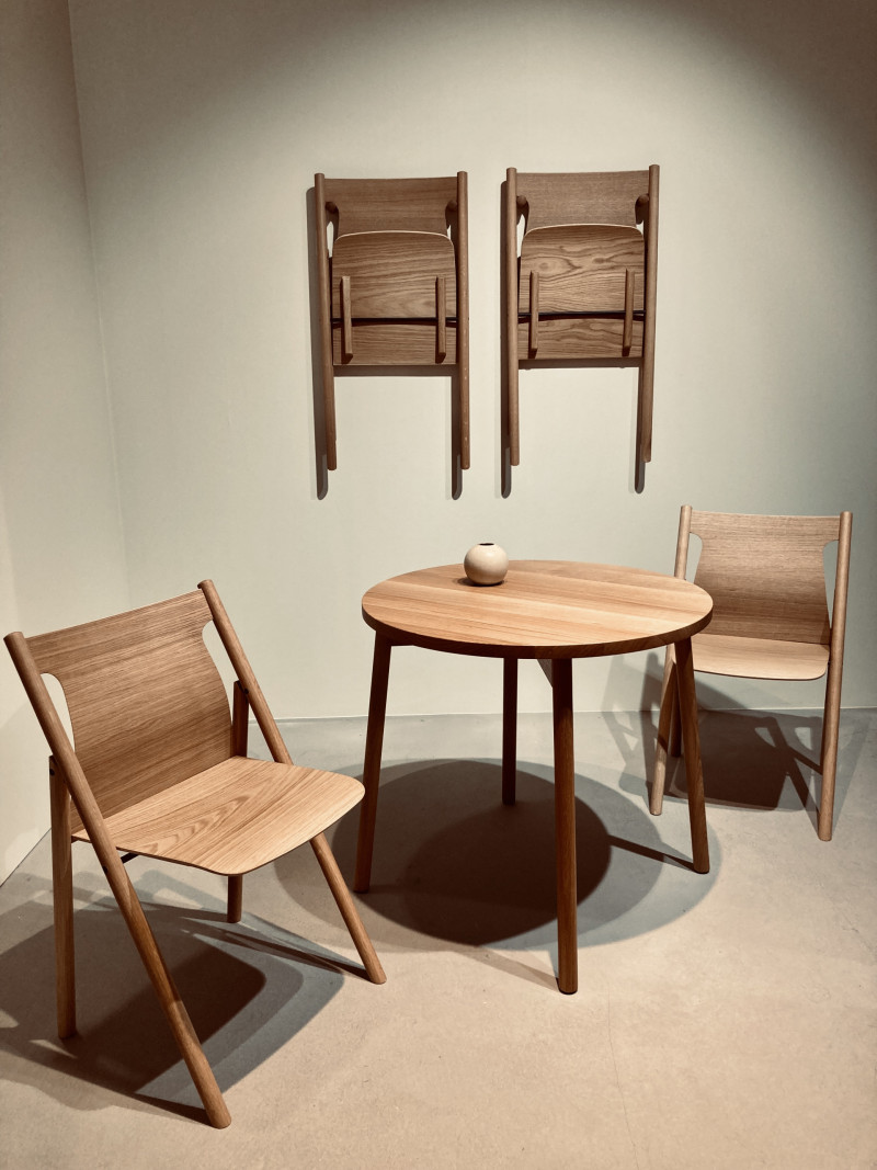 runder Tisch und 2 Klappstühle aus Nussbaumholz stehen in einem hellen Raum, 2 Klappstühle hängen auf gleicher Höhe an der Wand, kleine beige, kugelförmige Keramikvase steht auf dem Tisch