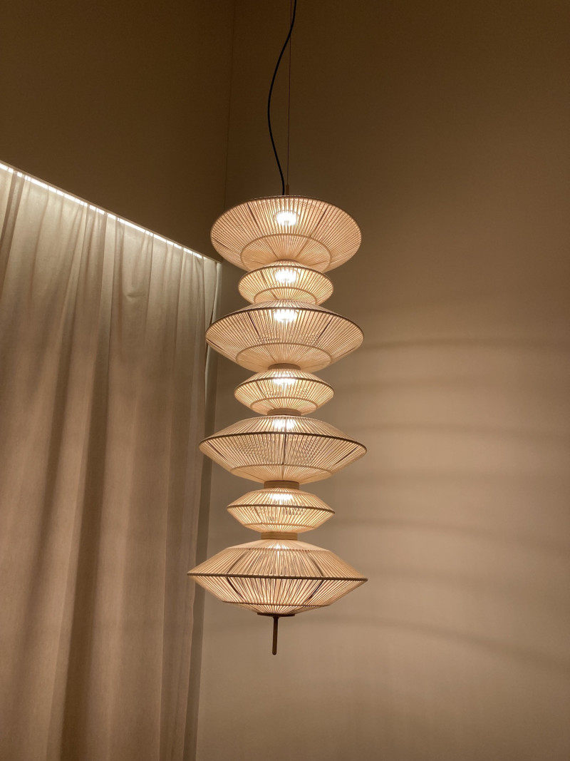 Lampe aus 7 runden, unterschiedlich großen Körpern, die mit Bast umwickelt sind, vor einer sandfarbenen Wand, links der Lampe ein heller Leinenvorhang