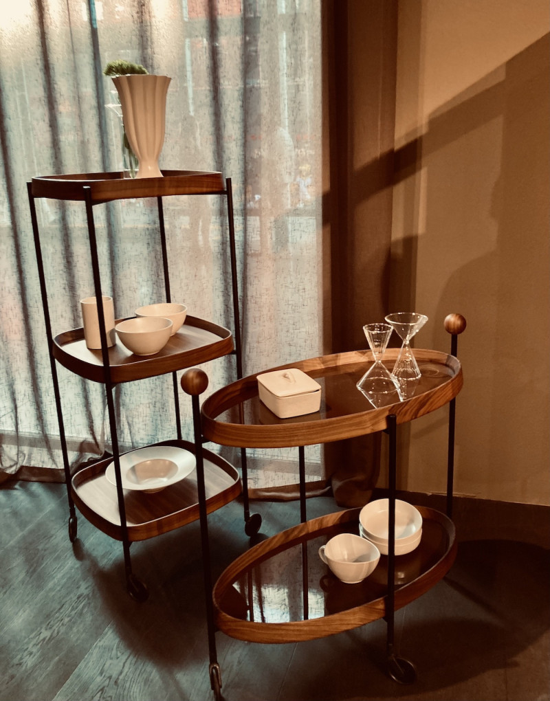 1 ovaler Teewagen aus Nussbaumholz mit 2 Böden, vor einer zimtfarbenen wand, darauf 1Tasse, 2 Schalen und 1 Dose aus heller Keramik und 2 Gläser, links daneben quadratisches Regal auf Rädern, mit 3 Böden und abgerundeten Ecken, vor einem hellen Leinenvorhang, darauf 1 Vase, 1 Becher, 2 Schalen und 1 tiefer Teller aus heller Keramik