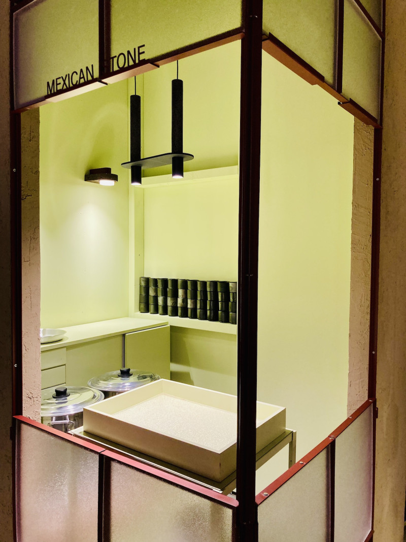 Immitation einer Garküche aus einer rotbraunen Metallkonstruktion und mattiertem Glas mit 2 großen rechtwinklig zueinander stehenden Fenster, pistaziengrünen Rückwänden und Mobiliar, 2 Töpfe, viele kleine schwarze Keramikschalen, 1 schwarze Wandleuchte, eine schwarze Pendelleuchte