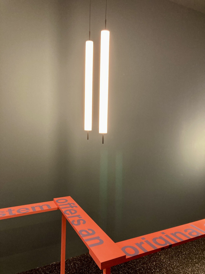 2 Stableuchten von der Decke hängend in gleicher Höhe, vor grauer Wand, darunter 3 Holzplanken, rechtwinklig angeordnet ähnlich einem Z, orange gestrichen und bedruckt auf Holzfüßen