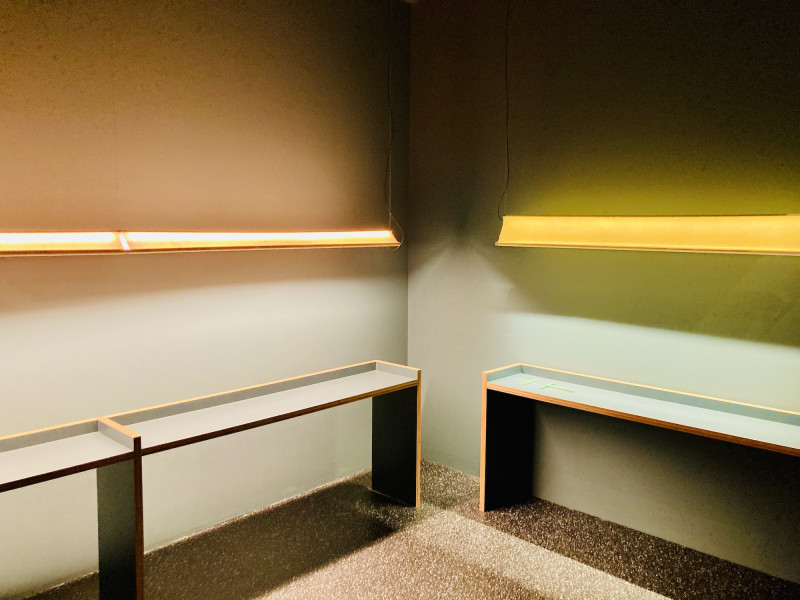 2 lange Tischleuchten in zartem Orange hängen einem gleichlangen, schmalen grüngrauen Tisch, vor gleichfarbiger Wand, in rechtem Winkel dazu, 1 gelbe Tischleuchte, über einem Tisch, gleiche Ausführung, vor gleicher Wand, Steinboden