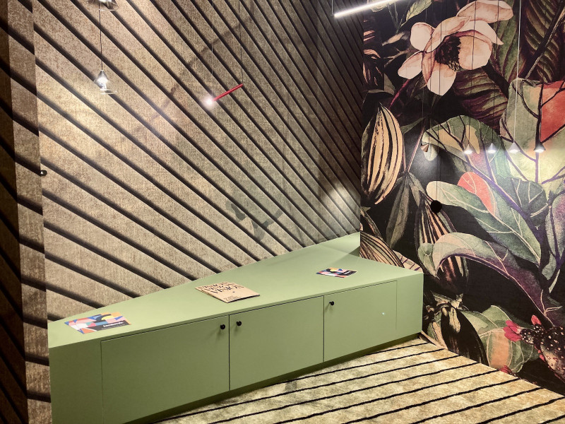 Raumecke mit lindgrünem, sich verjüngendem Sideboard, auf gelbem Teppich mit schwarzen Streifen, vor einer Tapete mit diagonal verlaufenden Planken, die rechtwinklig dazu angeordnete Wand hat eine schwarzgrundige Tapete mit grünen, beigen und roten Blättern und einer großen rosafarbenen geöffneten Blüte